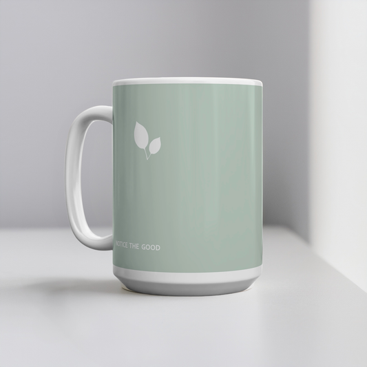 Green ceramic mug with small white leaf cluster design. Small white font at the bottom ‘Notice the good’. The mug sits on a white table, minimalist background.