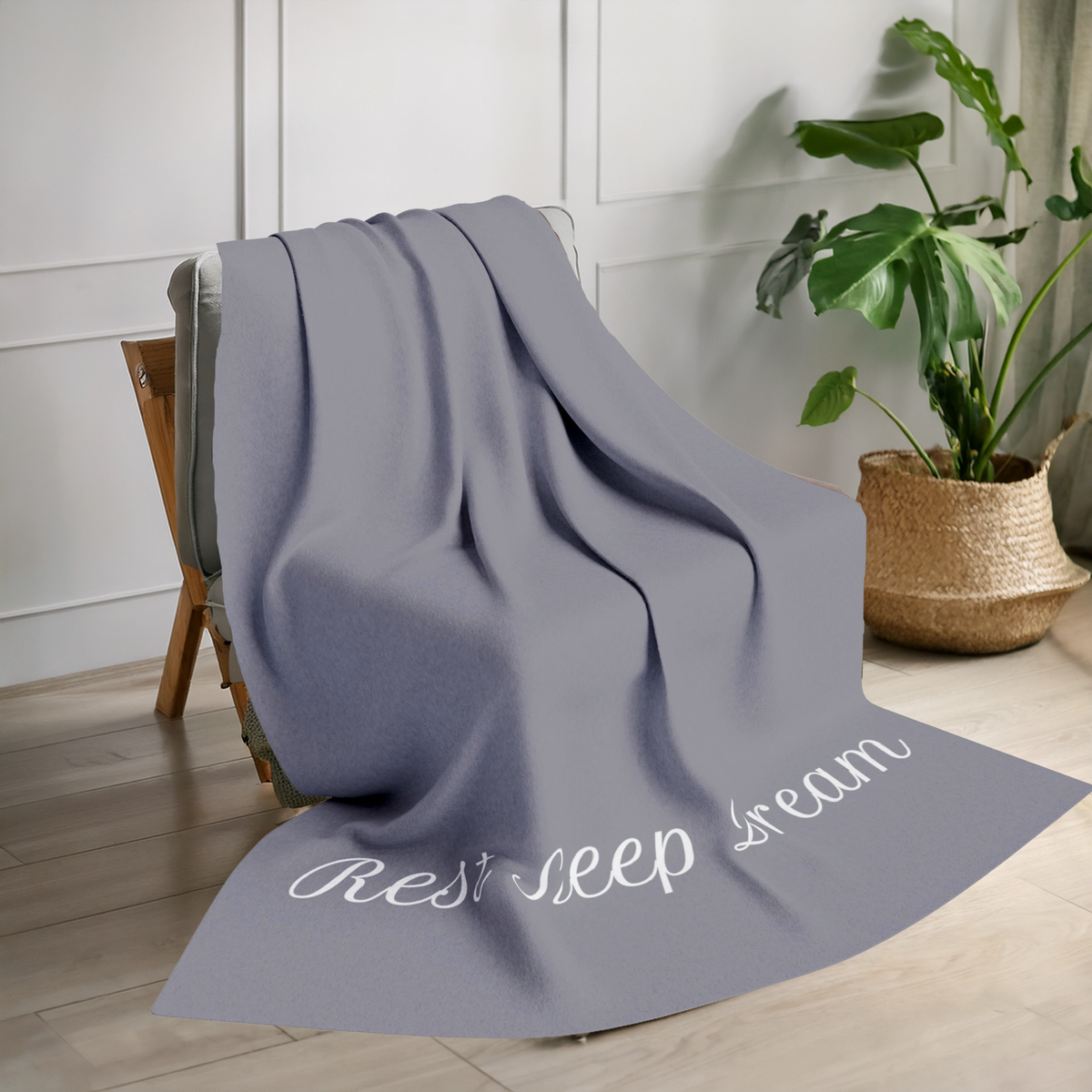 Dark grey blanket with large, elegant font in white for the text ‘Rest Sleep Dream’ along the bottom.  The blanket is on a chair in a living room with plant in the background.
