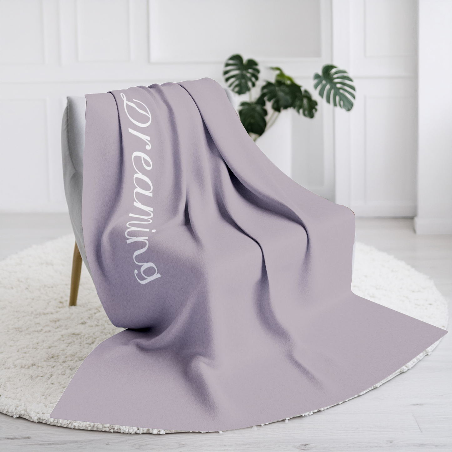 Light brown blanket with large, elegant font in white for the text ‘Dreaming'.  The blanket is on a chair in a living room with  plant in the background.