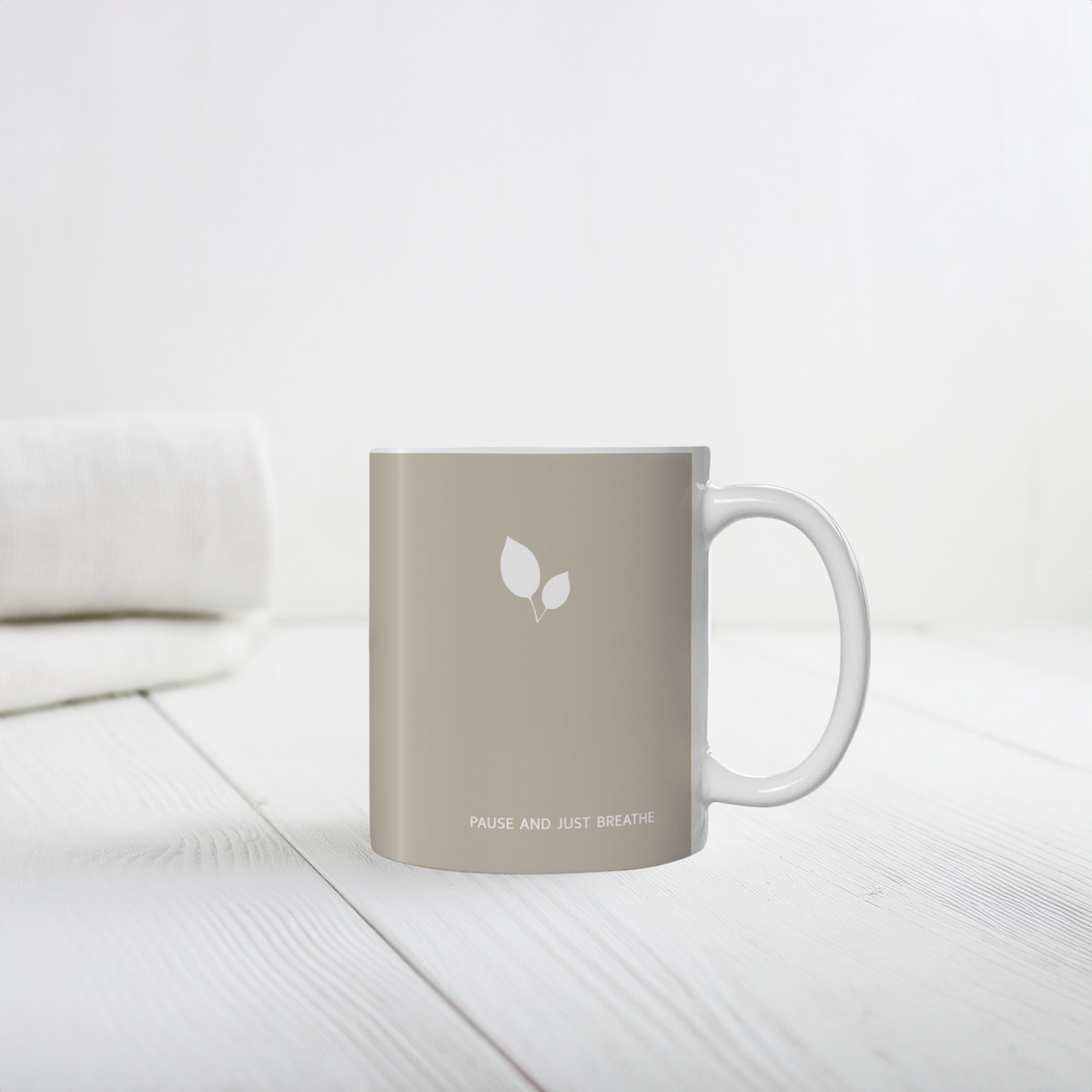 Brown ceramic mug with small white leaf cluster design. Small white font at the bottom ‘Pause and just breathe’. The mug sits on a white wooden table, minimalist background.
