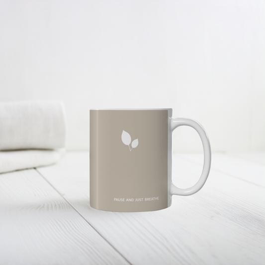 Brown ceramic mug with small white leaf cluster design. Small white font at the bottom ‘Pause and just breathe’. The mug sits on a white wooden table, minimalist background.