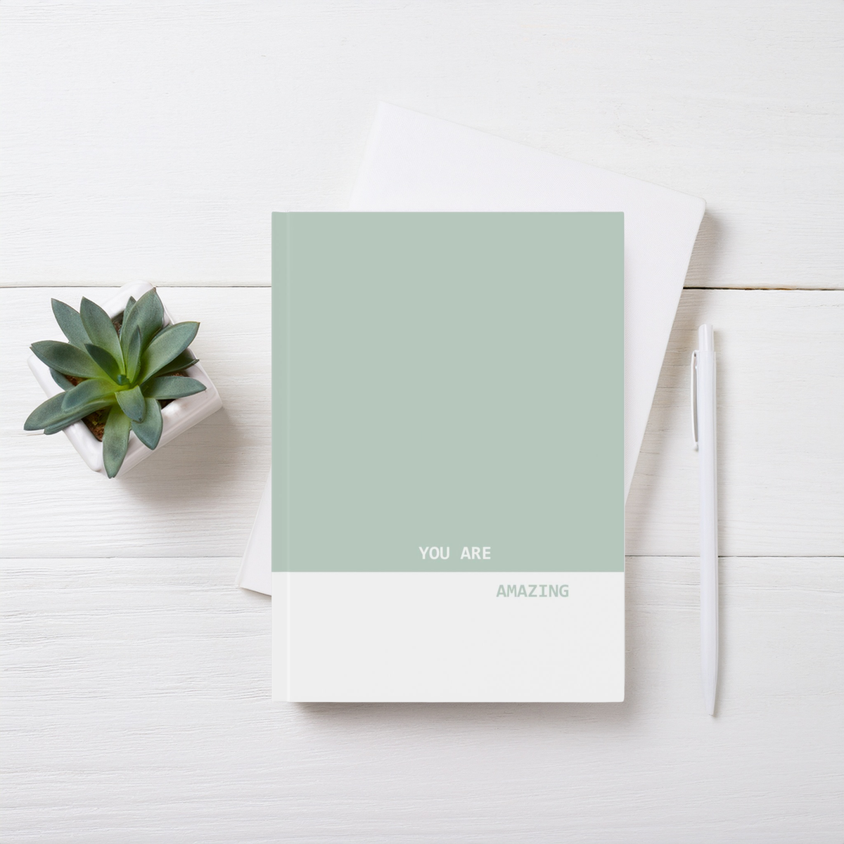 A5 hardcover journal notebook with a sage green top half and white bottom half reading 'YOU ARE AMAZING', styled on a white wood background with a small succulent and a white pen.