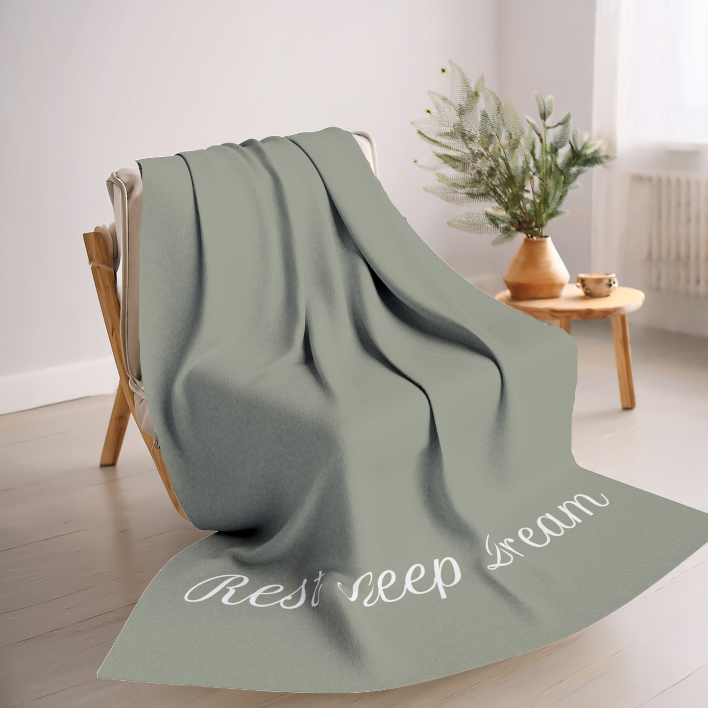 Green blanket with large, elegant font in white for the text ‘Rest Sleep Dream’ along the bottom.  The blanket is on a chair in a living room with small table and plant in the background.