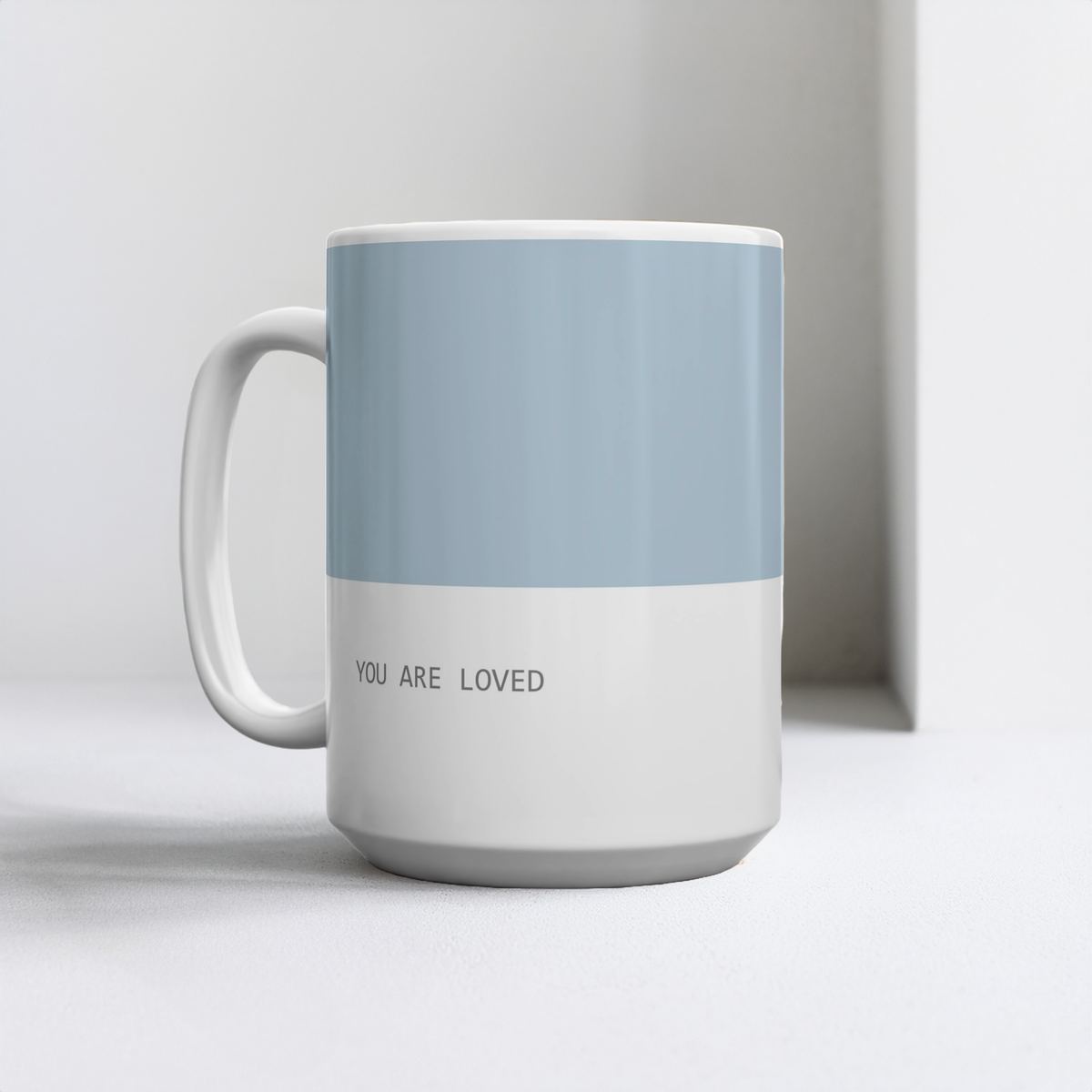 Blue and white ceramic mug with small font at the bottom ‘you are loved’. The mug sits on a white table, minimalist background.