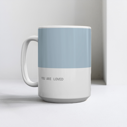 Blue and white ceramic mug with small font at the bottom ‘you are loved’. The mug sits on a white table, minimalist background.