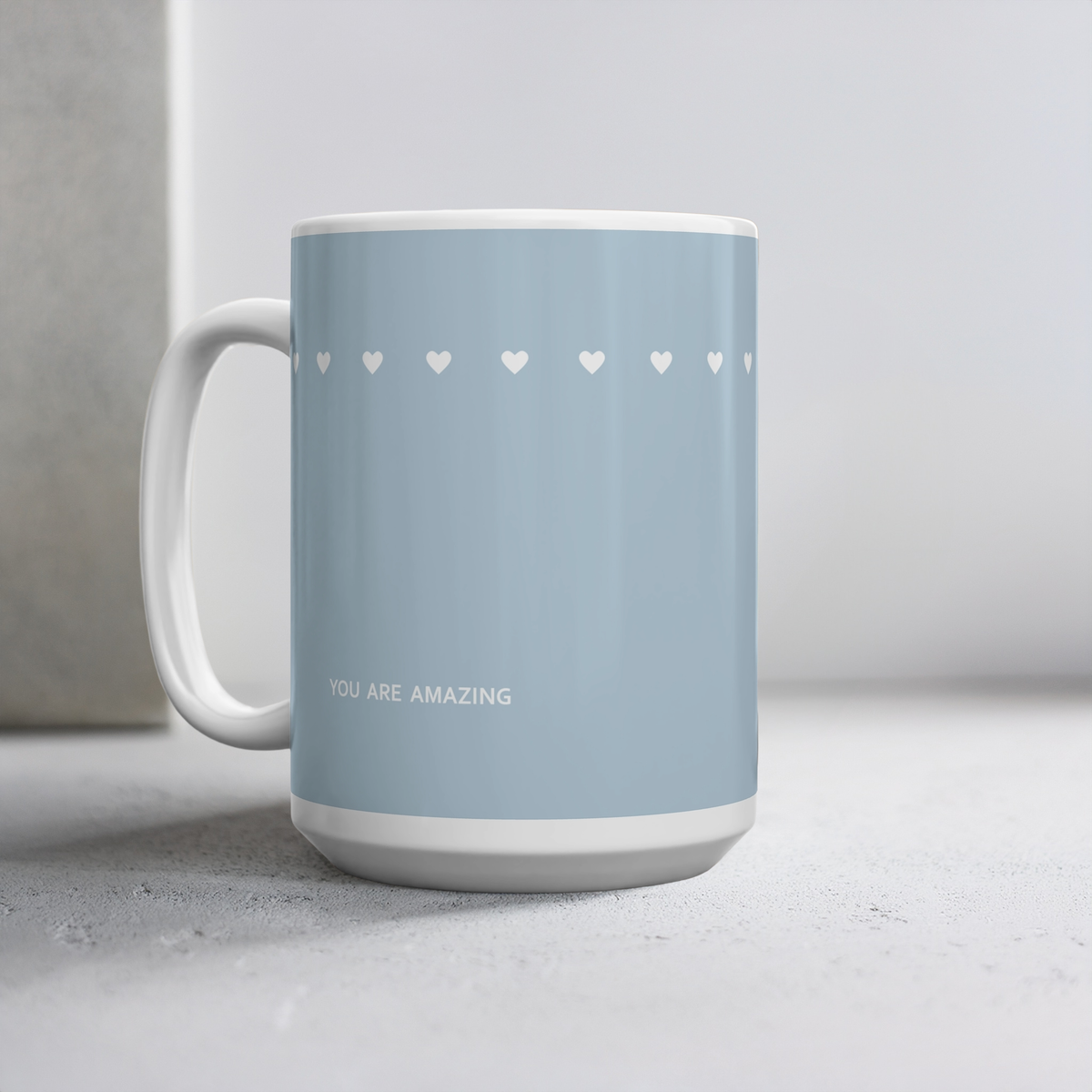 15oz blue ceramic mug with row of tiny white hearts. Small white font at the bottom ‘You are amazing’. The mug sits on a cement style surface, minimalist background.