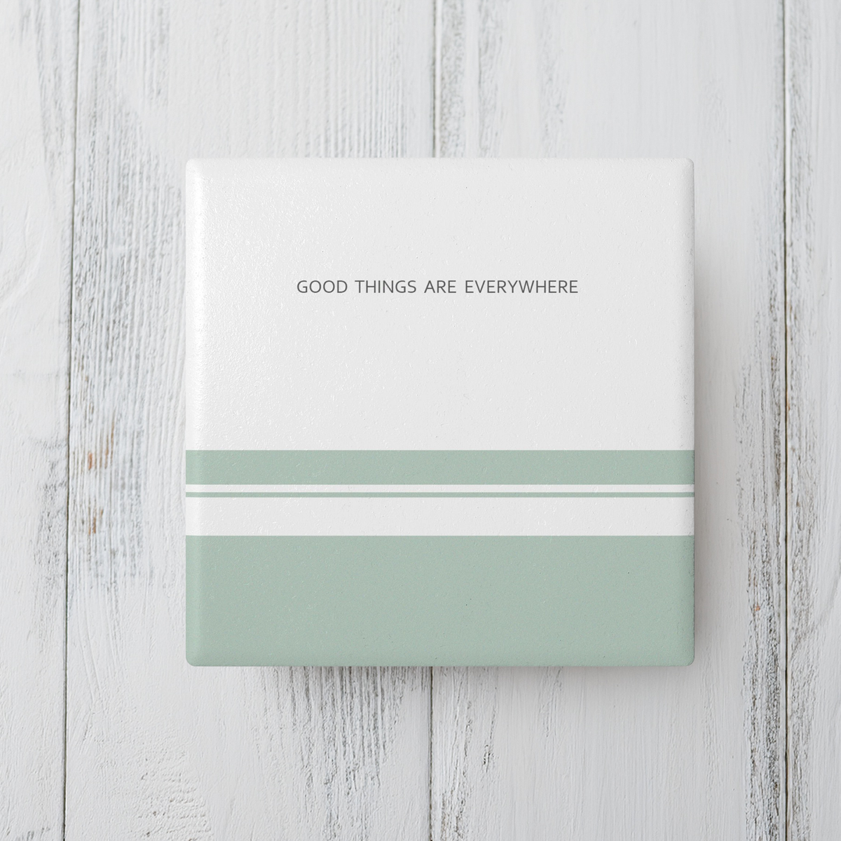 White square ceramic coaster with three sage green stripes in varying sizes at the bottom. The text 'Good things are everywhere' in dark grey small text at the top. The coaster sits on grey wooden surface.