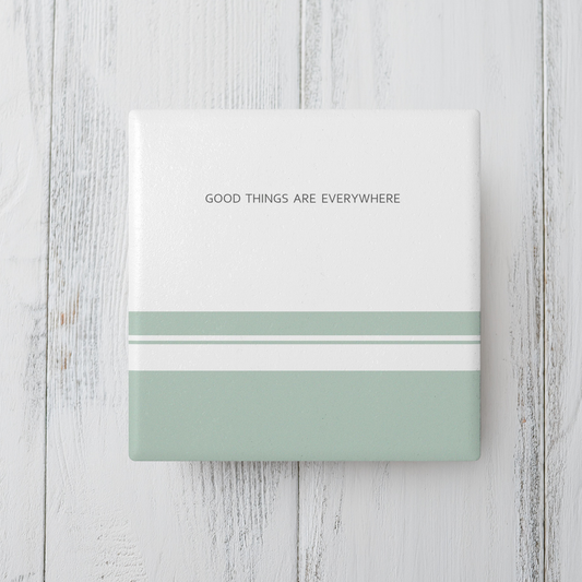 White square ceramic coaster with three sage green stripes in varying sizes at the bottom. The text 'Good things are everywhere' in dark grey small text at the top. The coaster sits on grey wooden surface.