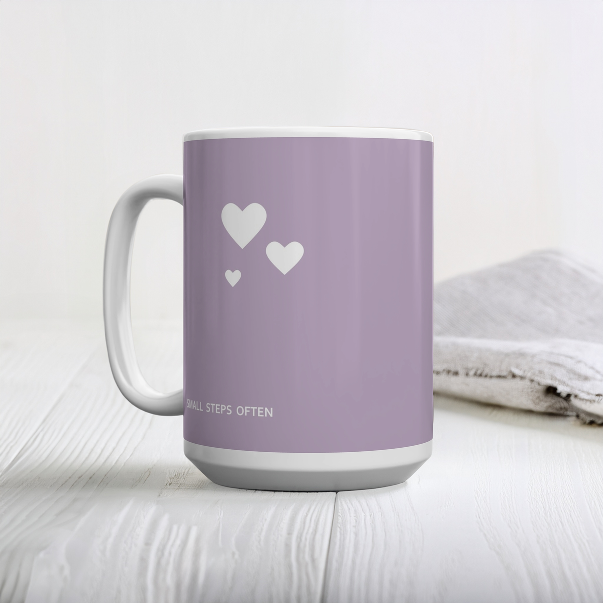 Purple ceramic mug with cluster of three white hearts. Small white font at the bottom ‘Small steps often’. The mug sits on a white wooden table, next to linen