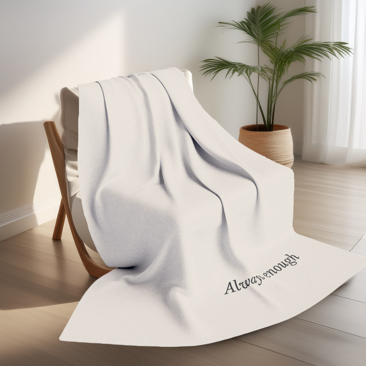 A cream beige throw blanket draped over a wooden chair in a bright room, with the phrase 'Always enough' printed in black script near one corner. A potted plant and sheer curtains are visible in the background.
