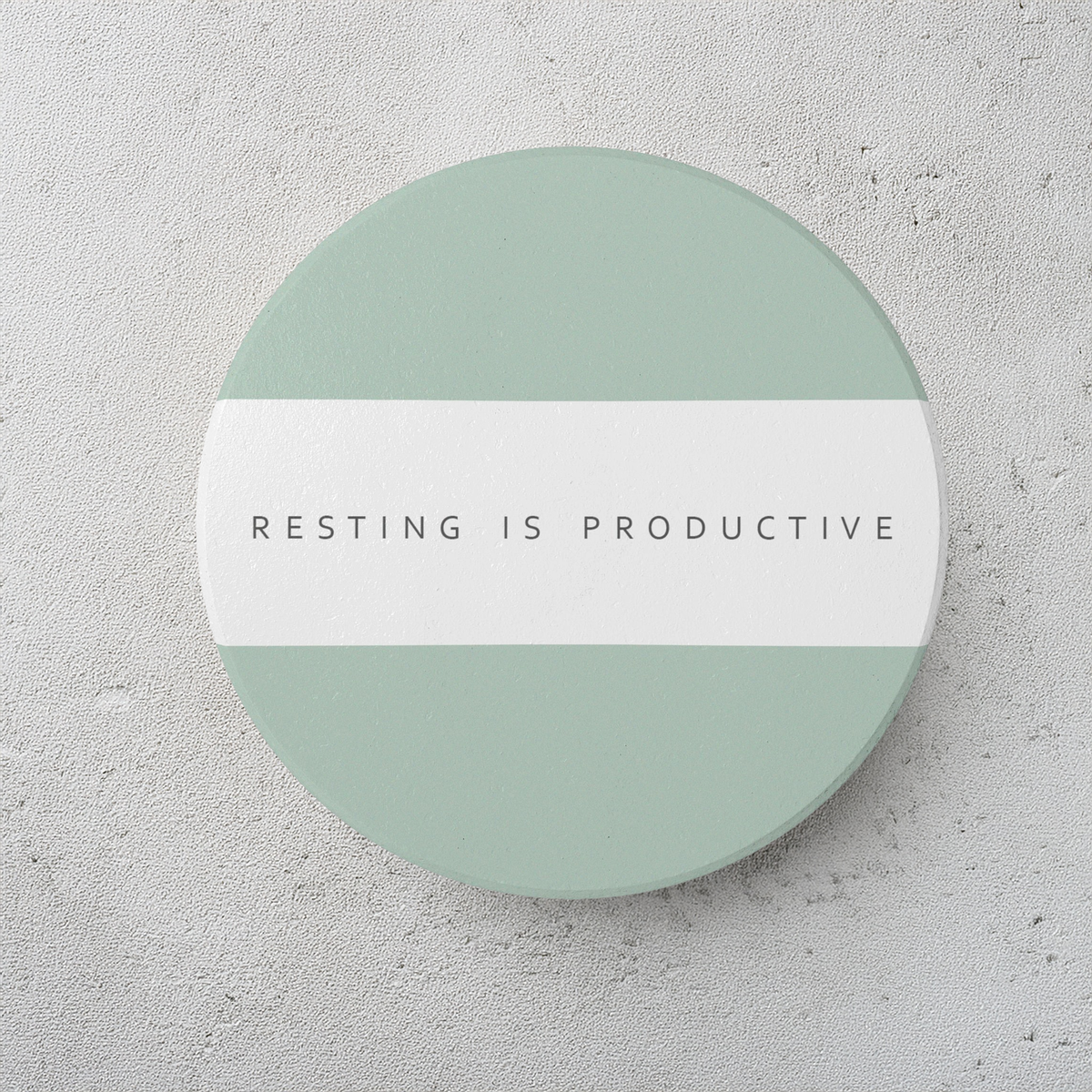 A round ceramic coaster with sage green top and bottom half and a white horizontal band across the center, featuring the text 'RESTING IS PRODUCTIVE' in dark gray. The coaster is on a textured light gray surface.