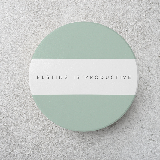 A round ceramic coaster with sage green top and bottom half and a white horizontal band across the center, featuring the text 'RESTING IS PRODUCTIVE' in dark gray. The coaster is on a textured light gray surface.