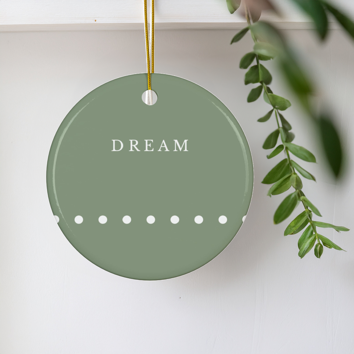 A round sage green ceramic Christmas ornament hanging from a gold string, featuring the word 'DREAM' in white serif letters and a row of white dots along the bottom, set against a white wall with a green plant branch on the right.