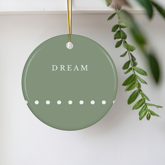 A round sage green ceramic Christmas ornament hanging from a gold string, featuring the word 'DREAM' in white serif letters and a row of white dots along the bottom, set against a white wall with a green plant branch on the right.