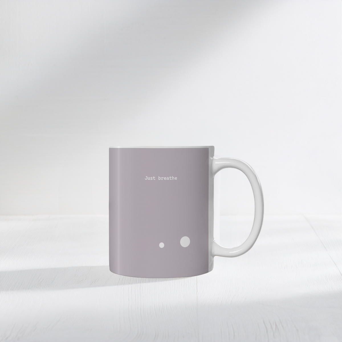 Taupe brown ceramic mug with two small white circle dots design. Small white font at the top ‘Just breathe’. The mug sits on a white surface, minimalist background.