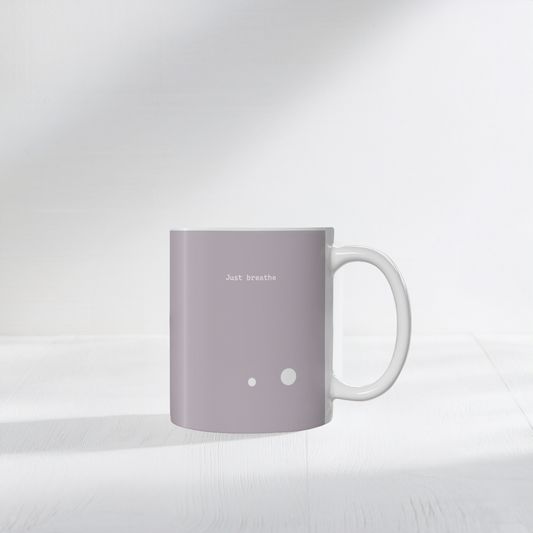 Taupe brown ceramic mug with two small white circle dots design. Small white font at the top ‘Just breathe’. The mug sits on a white surface, minimalist background.