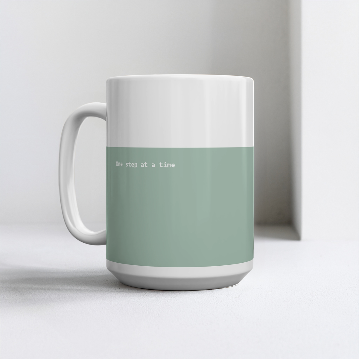 Green and white ceramic mug with small white font in the middle ‘One step at a time’. The mug sits on a white surface, minimalist background.