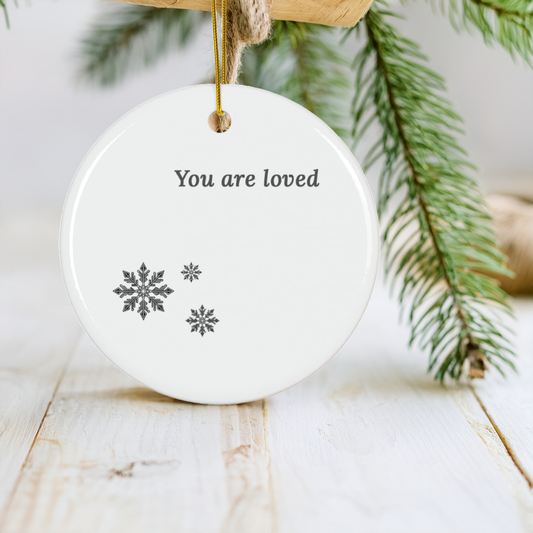 A round white ceramic Christmas ornament hanging from a wooden branch with jute twine, printed with the words 'You are loved' and three gray snowflakes, set against a soft-focus background of pine branches and a white wooden surface.