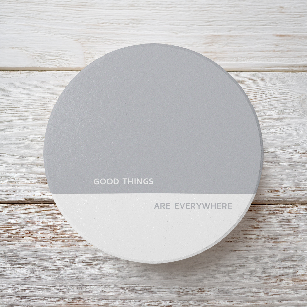 A round white and gray coaster with the printed text 'GOOD THINGS ARE EVERYWHERE' in gray, shown on a light whitewashed wood background.