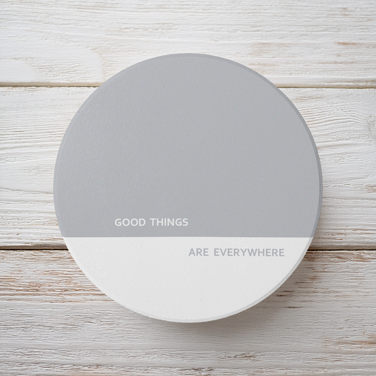A round white and gray coaster with the printed text 'GOOD THINGS ARE EVERYWHERE' in gray, shown on a light whitewashed wood background.