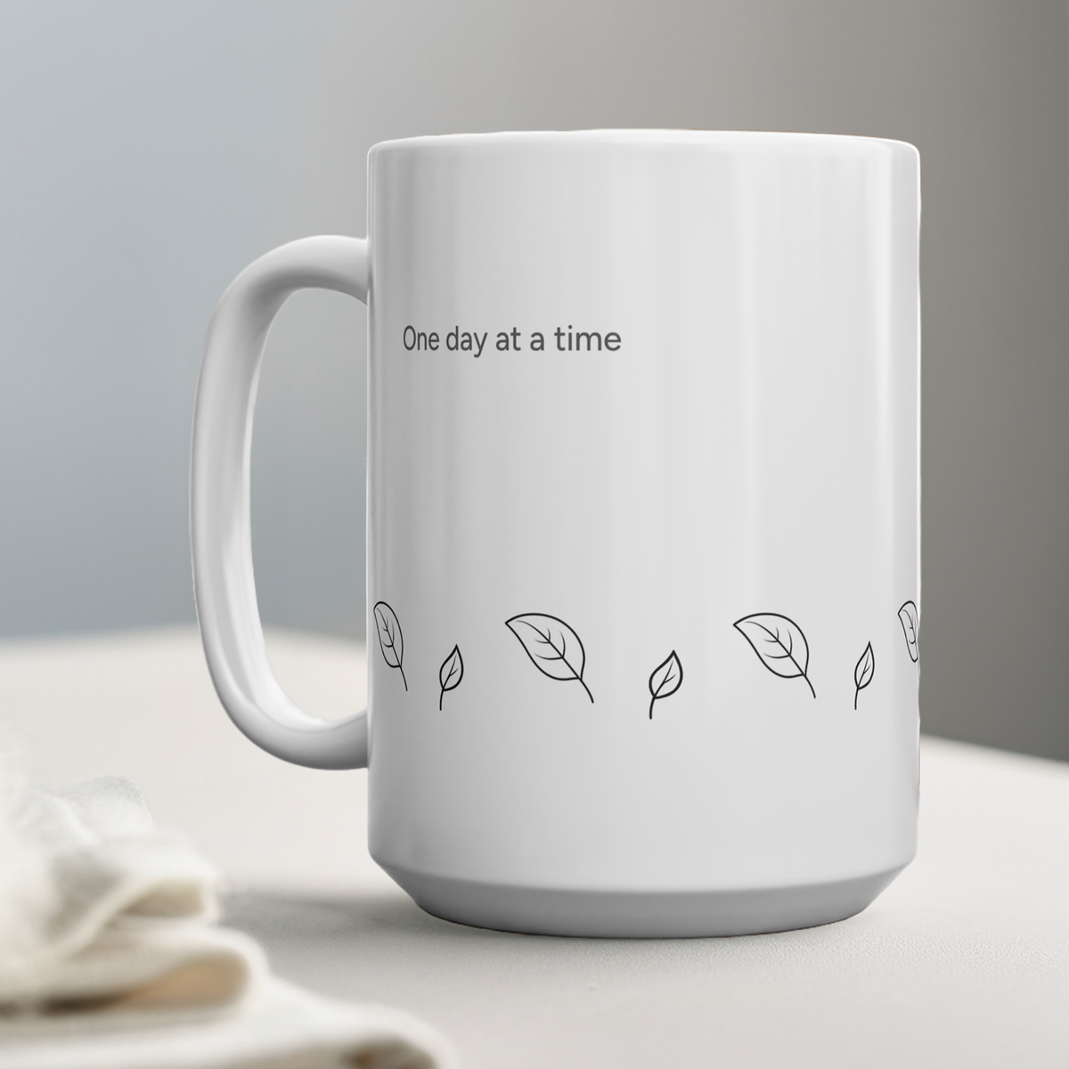 A white ceramic mug with the text 'One day at a time' and small leaf illustrations, shown on a light tabletop against a soft gray background.