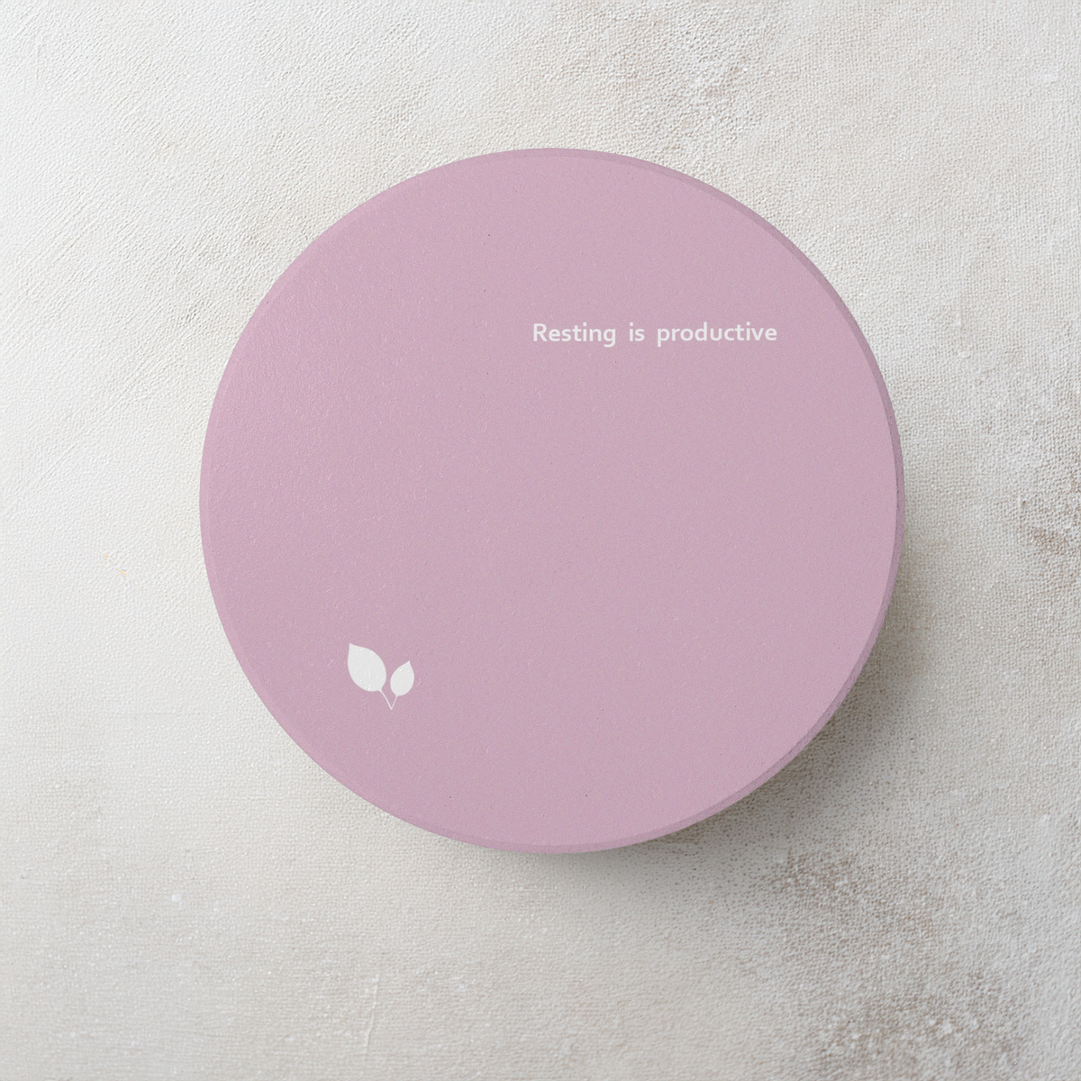 Pink round ceramic coaster with delicate leaf motif design, and small, modern style text ‘Resting is productive’ in white font. The coaster is on a cement style surface.