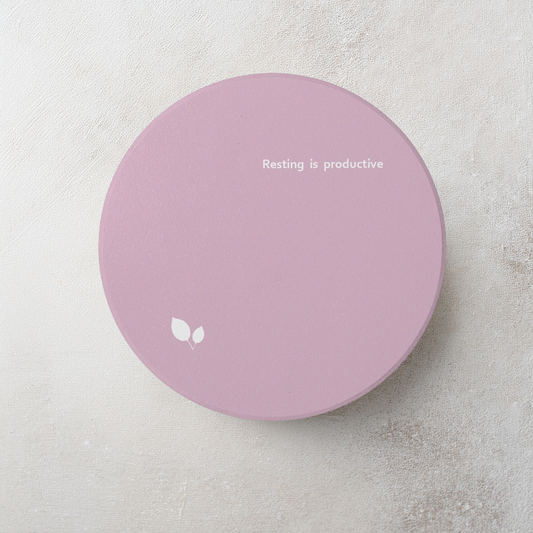 Pink round ceramic coaster with delicate leaf motif design, and small, modern style text ‘Resting is productive’ in white font. The coaster is on a cement style surface.