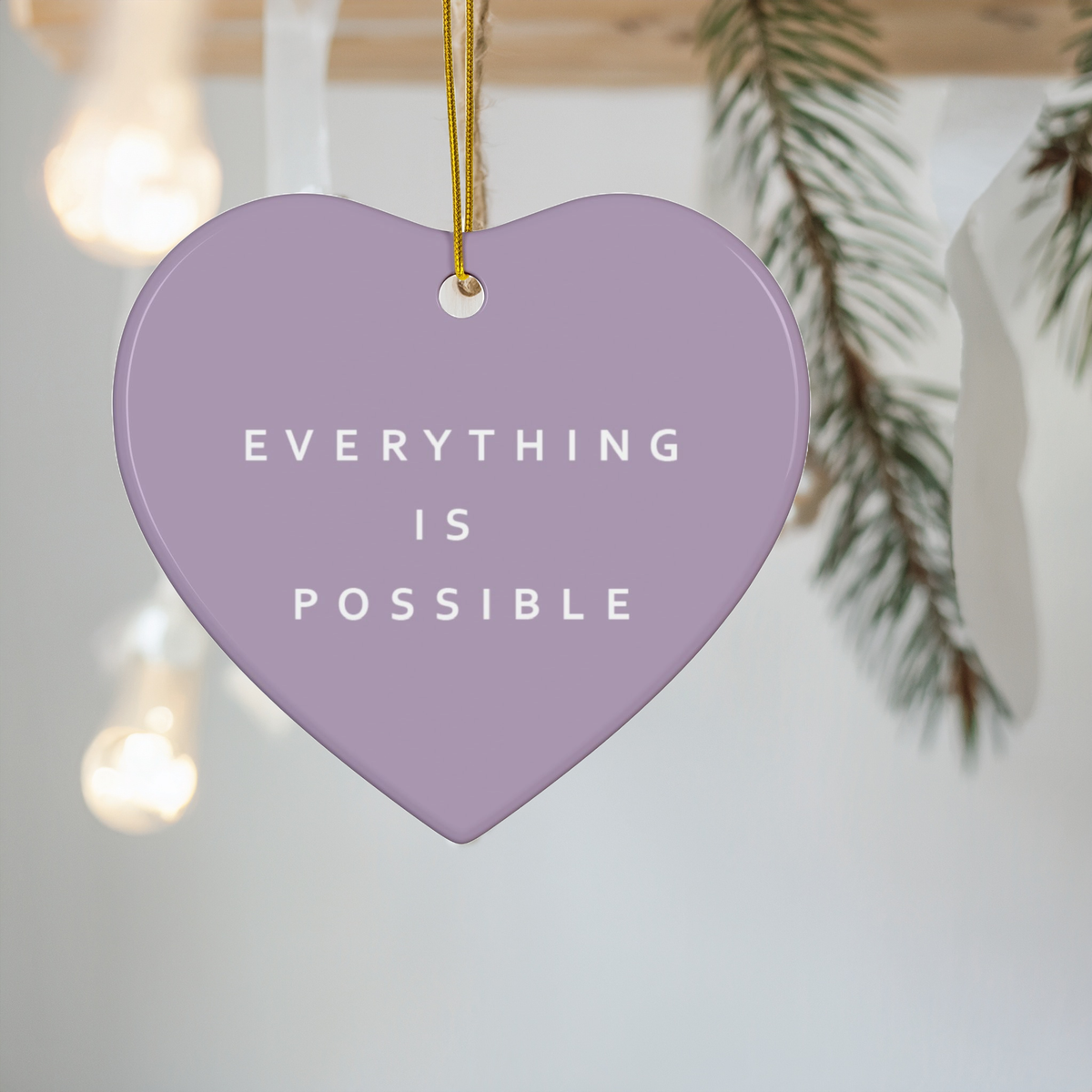 A pastel lavender heart-shaped hanging ornament with the phrase 'EVERYTHING IS POSSIBLE' in white text, suspended by a gold string against a softly blurred holiday background with lights and pine branches.