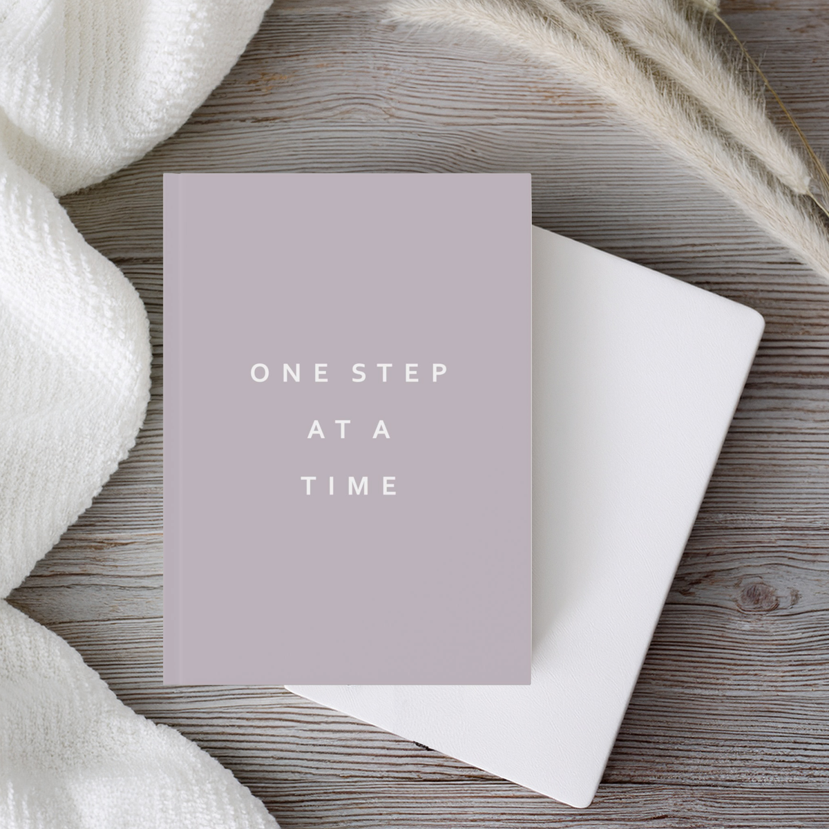 A5 hardcover. taupe brown notebook journal with the phrase 'ONE STEP AT A TIME' in white sans-serif text, placed on top of a plain white notebook on a rustic wood surface with a white knit fabric and dried grasses around it.