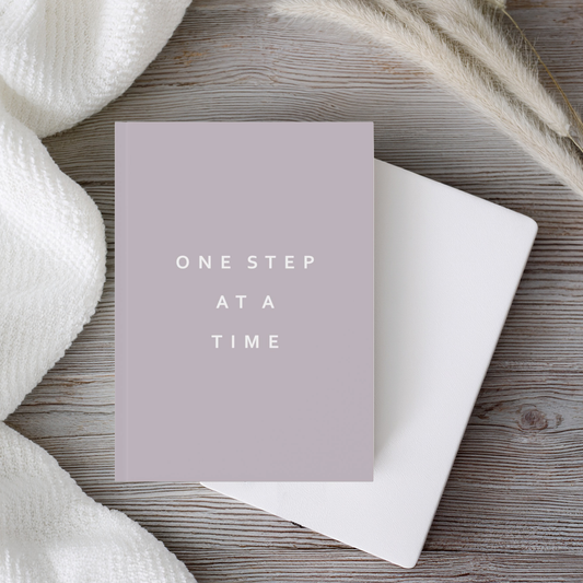 A5 hardcover. taupe brown notebook journal with the phrase 'ONE STEP AT A TIME' in white sans-serif text, placed on top of a plain white notebook on a rustic wood surface with a white knit fabric and dried grasses around it.