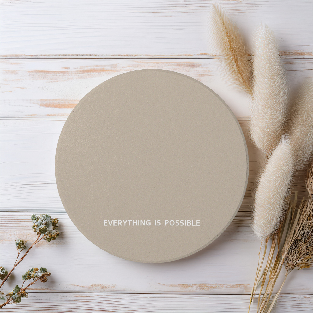 Beige round ceramic coaster with modern style text ‘Everything is possible’ in white font. The coaster is on a wooden surface beside wildflowers.
