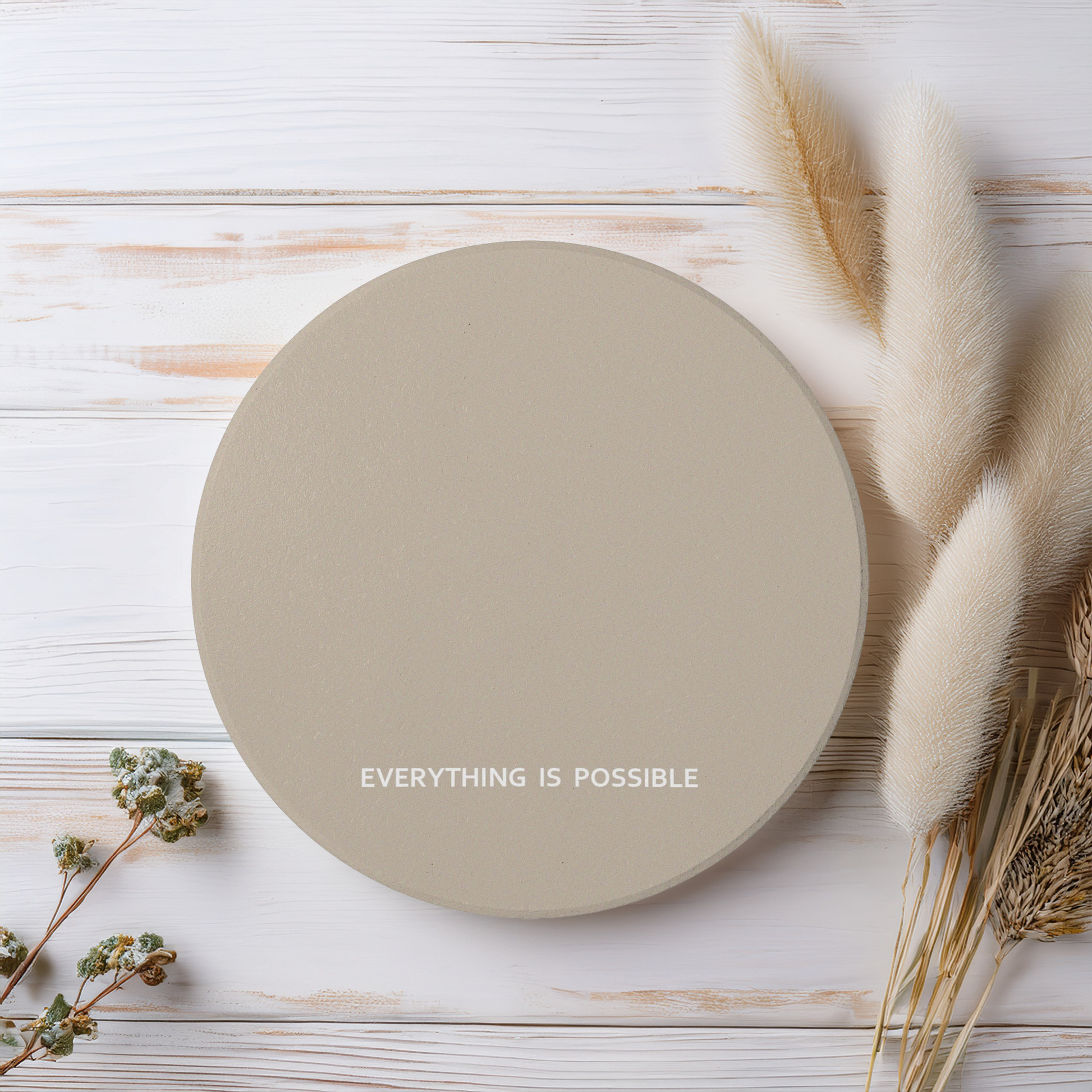 Beige round ceramic coaster with modern style text ‘Everything is possible’ in white font. The coaster is on a wooden surface beside wildflowers.