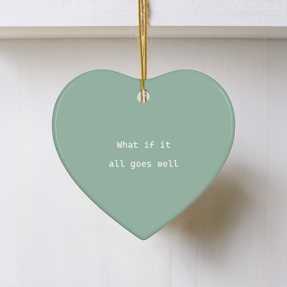 A heart-shaped ceramic ornament in a muted sage green color, hanging from a gold string. The ornament features the white printed text 'What if it all goes well' and is set against a clean white paneled background.