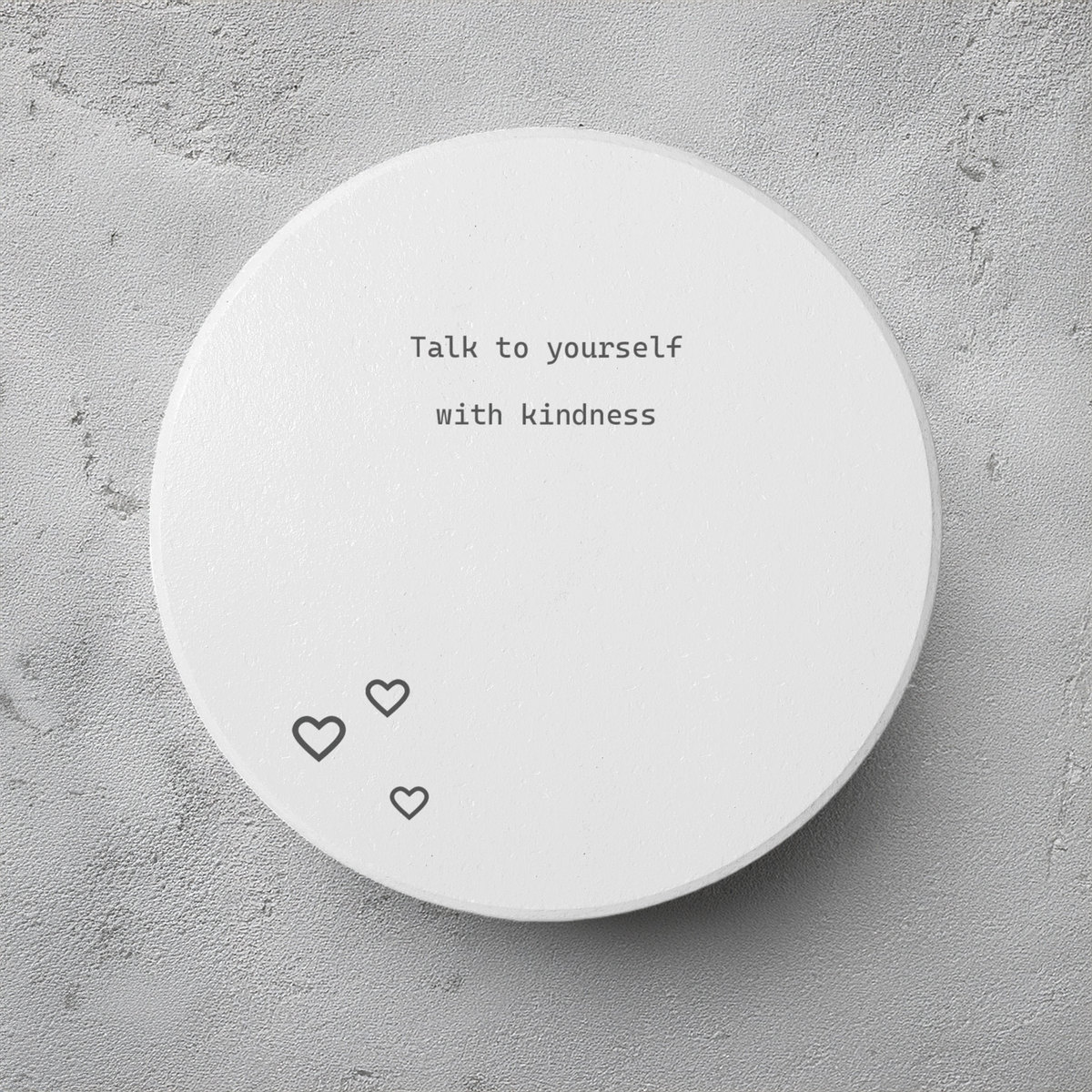 A round white coaster with the typewriter-style text 'What if it all goes well' printed in black, shown on a textured gray concrete background. The coaster has three small outlined hearts at the bottom.
