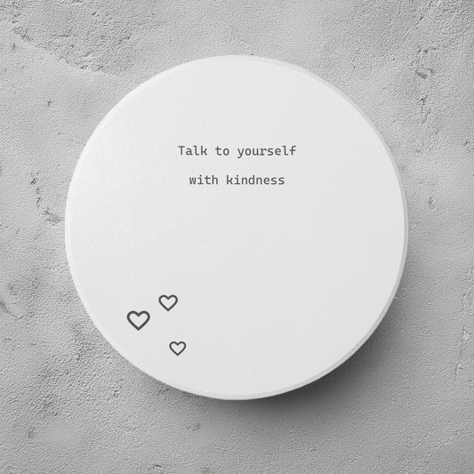 A round white coaster with the typewriter-style text 'What if it all goes well' printed in black, shown on a textured gray concrete background. The coaster has three small outlined hearts at the bottom.