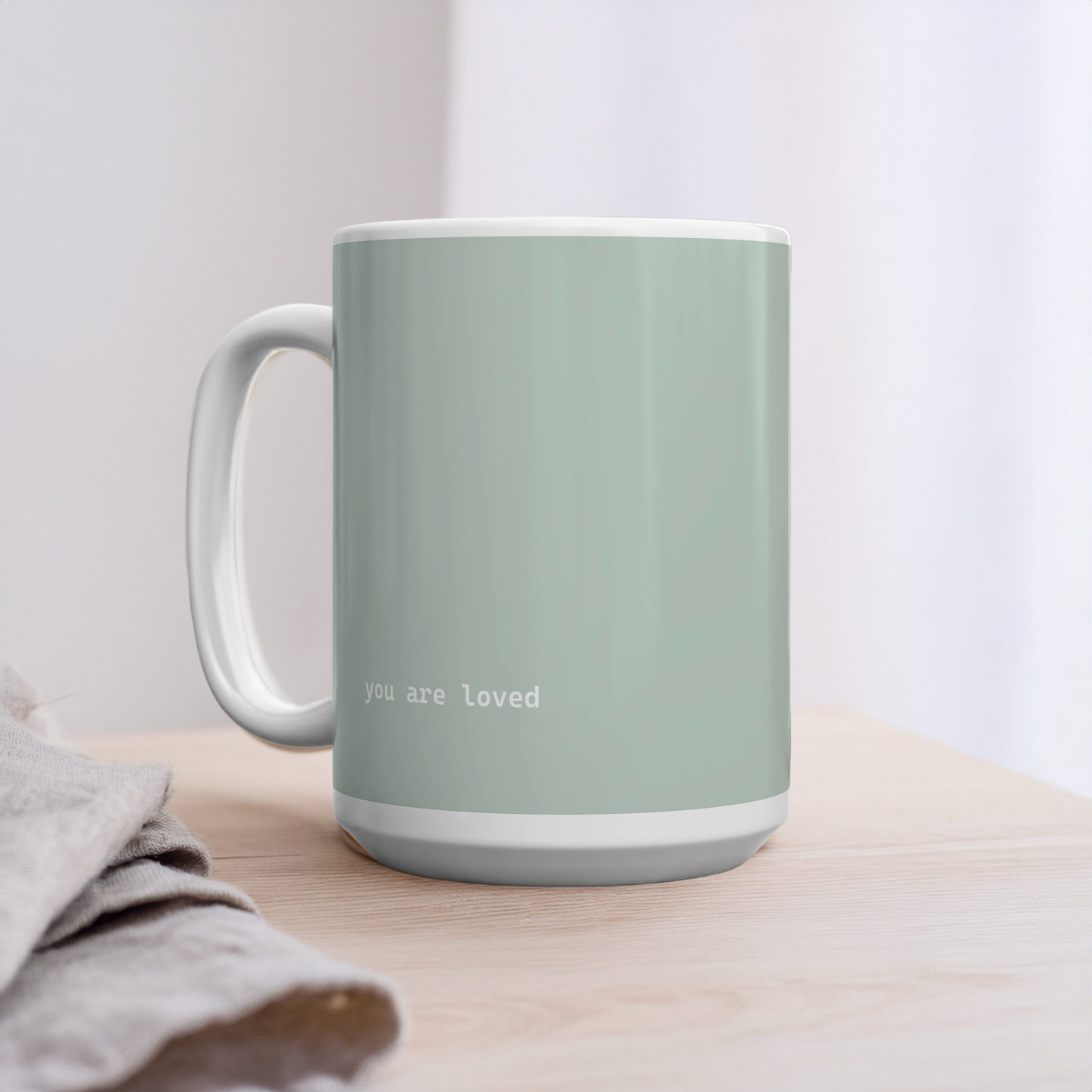Green ceramic mug with small white font at the bottom ‘you are loved’. The mug sits on a wooden table next to linens, minimalist background.