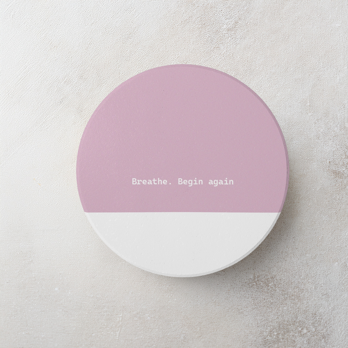 Pink and white round ceramic coaster with modern style text ‘Breathe. Begin again’ in white font. The coaster is on a cement style surface.
