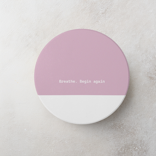 Pink and white round ceramic coaster with modern style text ‘Breathe. Begin again’ in white font. The coaster is on a cement style surface.