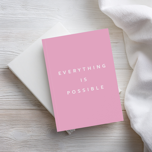 A pink hardcover notebook with the words 'EVERYTHING IS POSSIBLE' in white text, resting on a white notebook and a white textured fabric on a light wood surface.