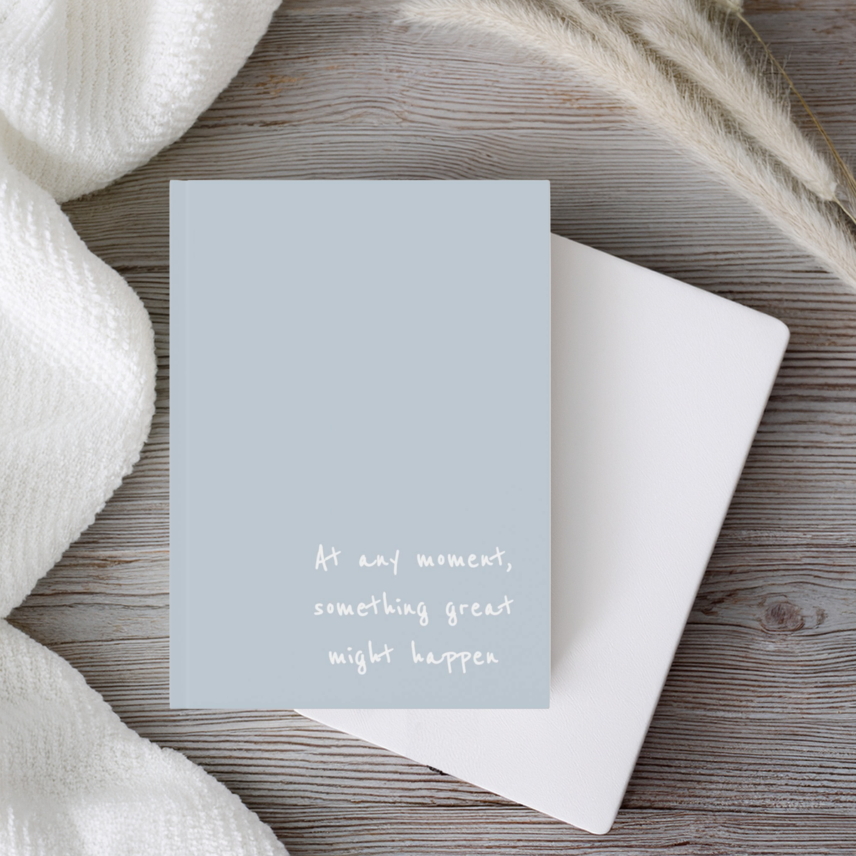 A light blue, hardcover, a5 journal notebook with white handwritten style text reading 'At any moment, something great might happen', styled on a rustic wood background with a white envelope and soft white textiles.