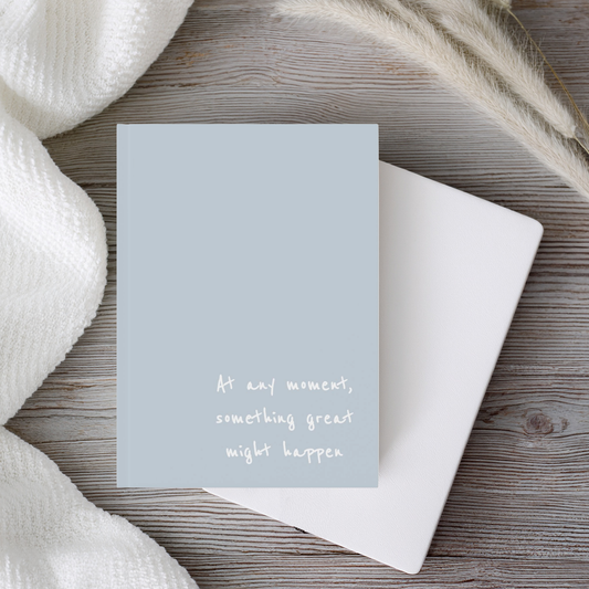 A light blue, hardcover, a5 journal notebook with white handwritten style text reading 'At any moment, something great might happen', styled on a rustic wood background with a white envelope and soft white textiles.