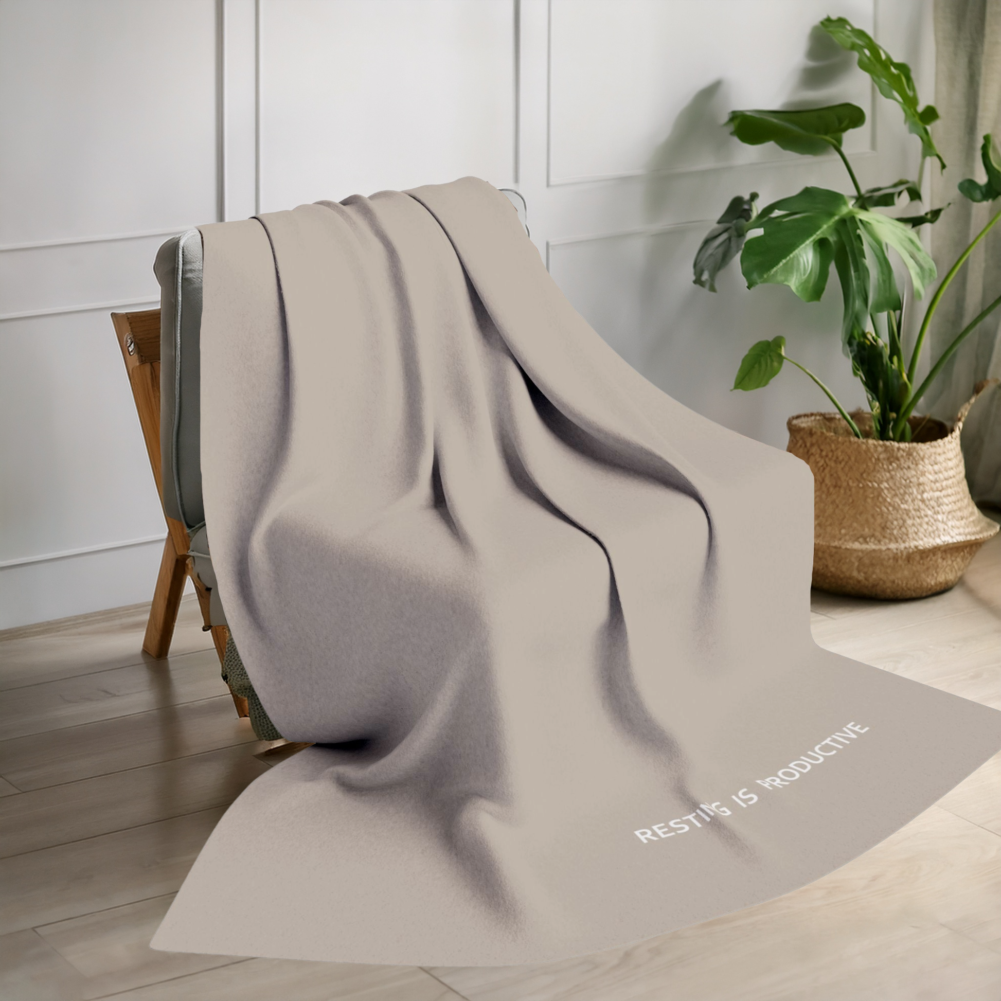 Beige blanket with tiny modern font in white for the text ‘Resting is Productive’ along the bottom. The blanket is on a chair in a living room with a plant in the background.