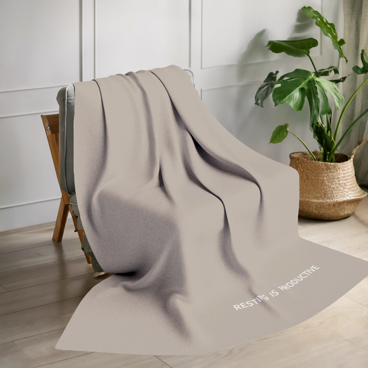 Beige blanket with tiny modern font in white for the text ‘Resting is Productive’ along the bottom. The blanket is on a chair in a living room with a plant in the background.