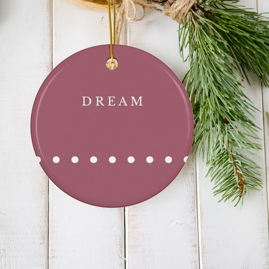 A round dark red ceramic Christmas ornament hanging from a gold string, featuring the word 'DREAM' in white serif letters and a row of white dots. The ornament is styled on white wooden planks next to green pine branches.