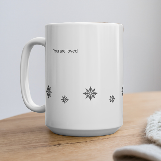 A white ceramic mug with the words 'You are loved' and small snowflake designs printed on it, sitting on a wooden surface.