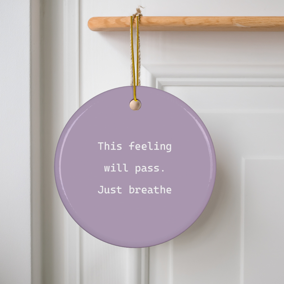 A round lavender purple ceramic ornament hanging from a wooden doorknob on a white paneled door. The ornament features the printed white text 'This feeling will pass.