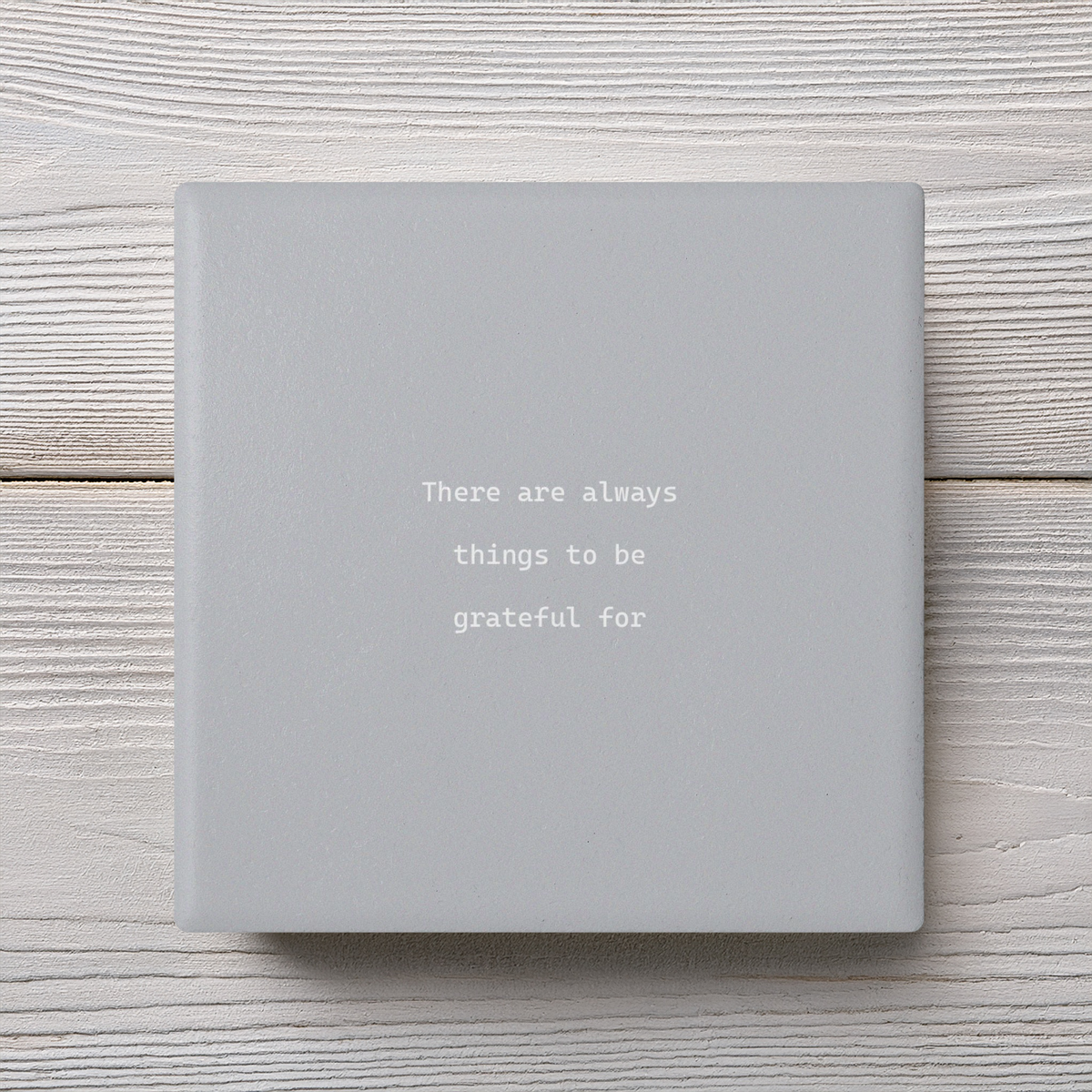 A square light gray ceramic coaster with the typographic quote 'There are always things to be grateful for' in white, displayed on a white wood plank background.