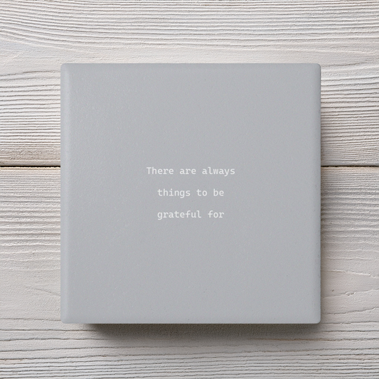 A square light gray ceramic coaster with the typographic quote 'There are always things to be grateful for' in white, displayed on a white wood plank background.