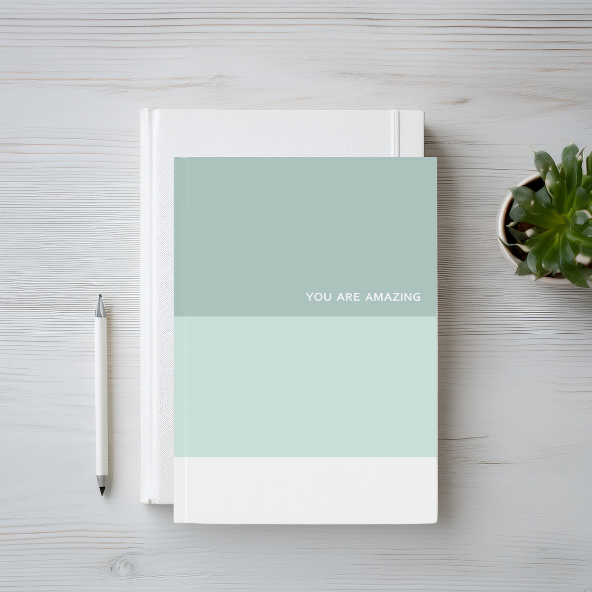A hardcover notebook with a minimalist color-blocked cover in sage green, mint, and white, featuring the text 'YOU ARE AMAZING' in white. The notebook is displayed on a light gray wood-grain desk alongside a silver pen and a small potted succulent.