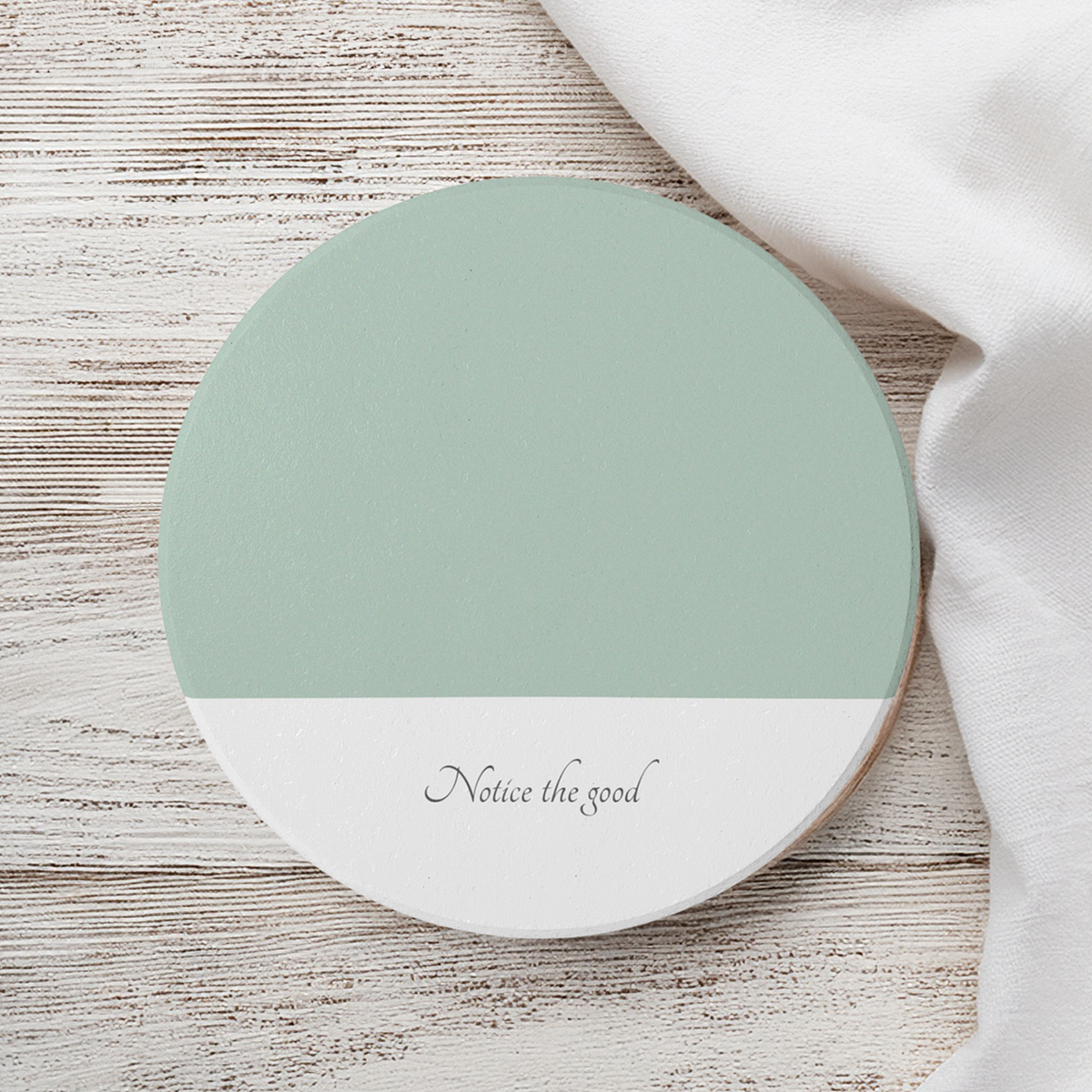 Green and white ceramic round coaster. The text 'Notice the good' in the bottom white part. The coaster sits on a wooden surface next to white linen.
